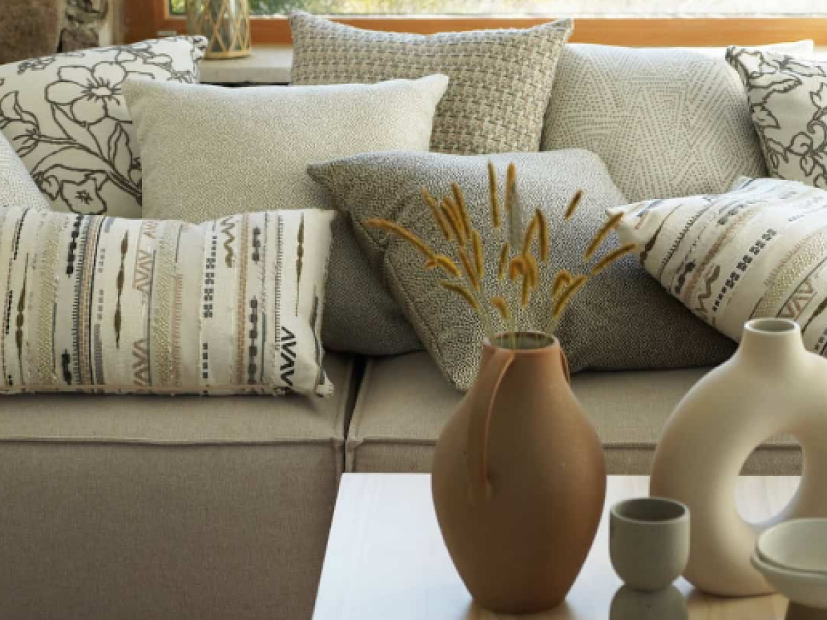 Verschiedene Kissen im Grundton Grau mit verschiedenen Mustern und aus verschiedenen Materialien liegen auf einem grauen Sofa. Davor steht eine braune Vase auf einem Tisch.